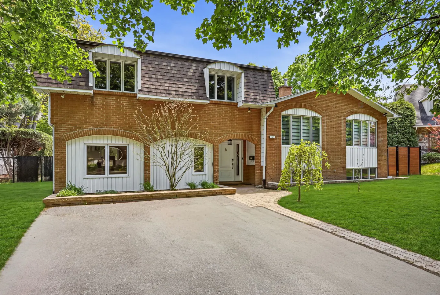 Brick house with a brown roof, white trim, and arched windows. A driveway leads to the front door, with trees framing the view.