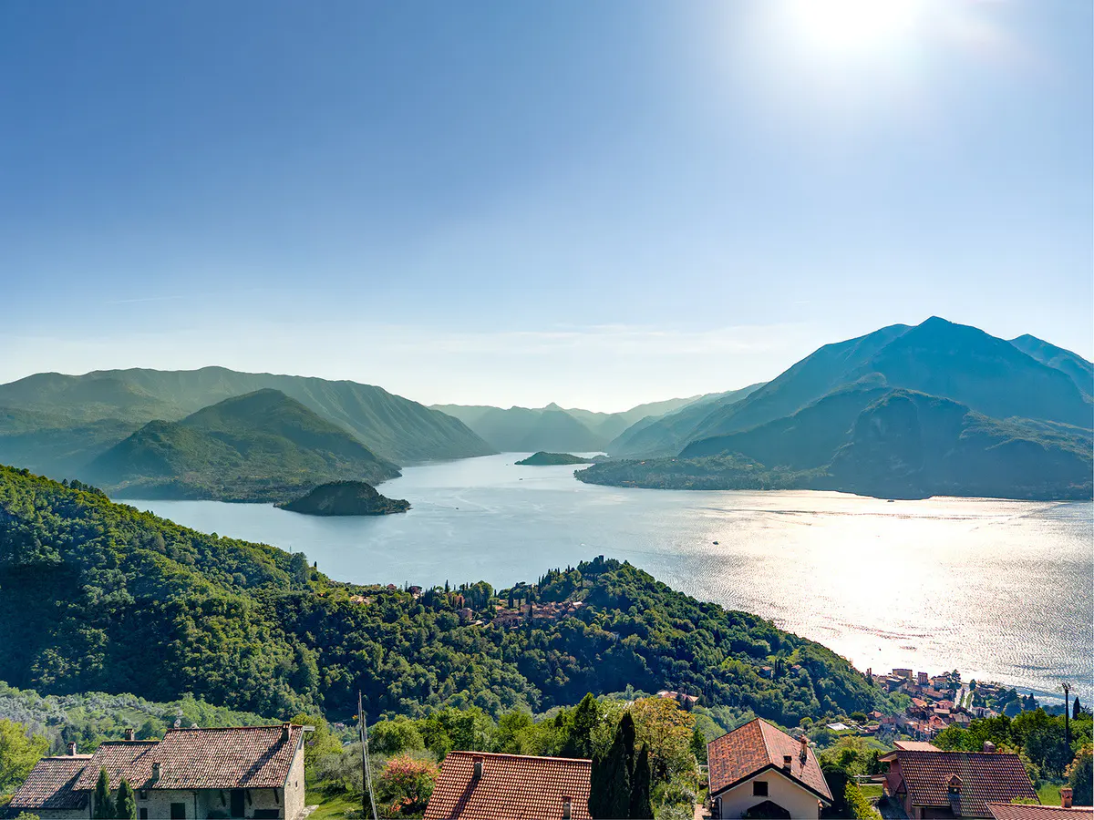 Scenic view of Lake Como, Italy, with green mountains, blue water reflecting sunlight, and houses with red tile roofs in the foreground.