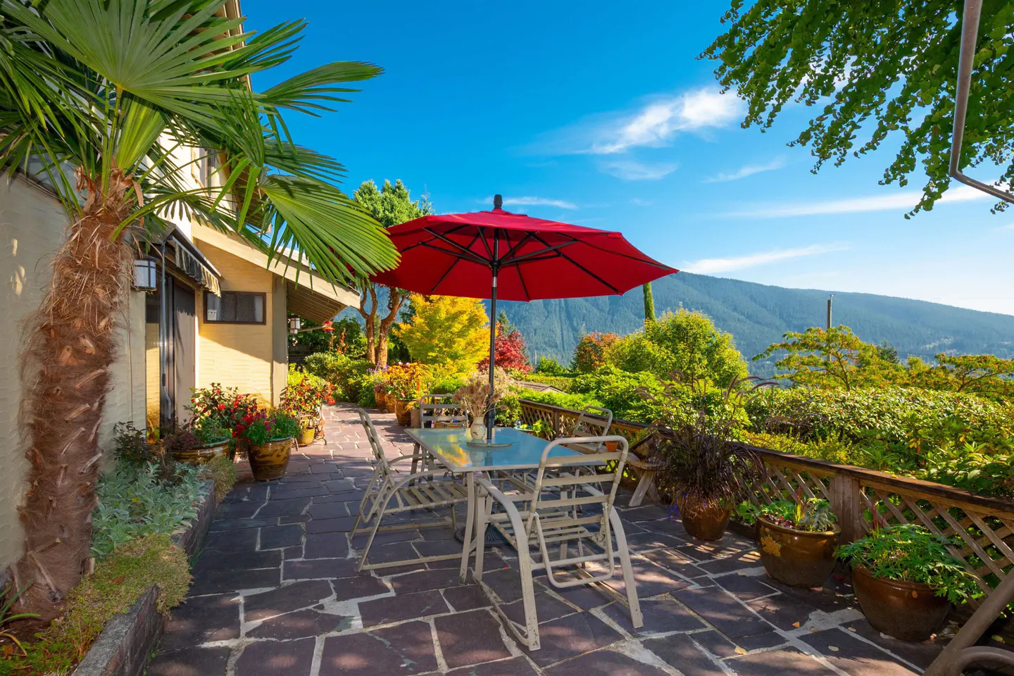 Outdoor patio with stone floor, table, chairs, and red umbrella. Lush greenery and mountain views in the background.