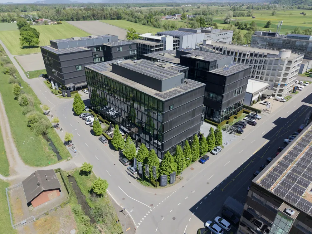 Aerial view of modern office buildings with solar panels, surrounded by green fields and roads with parked cars.