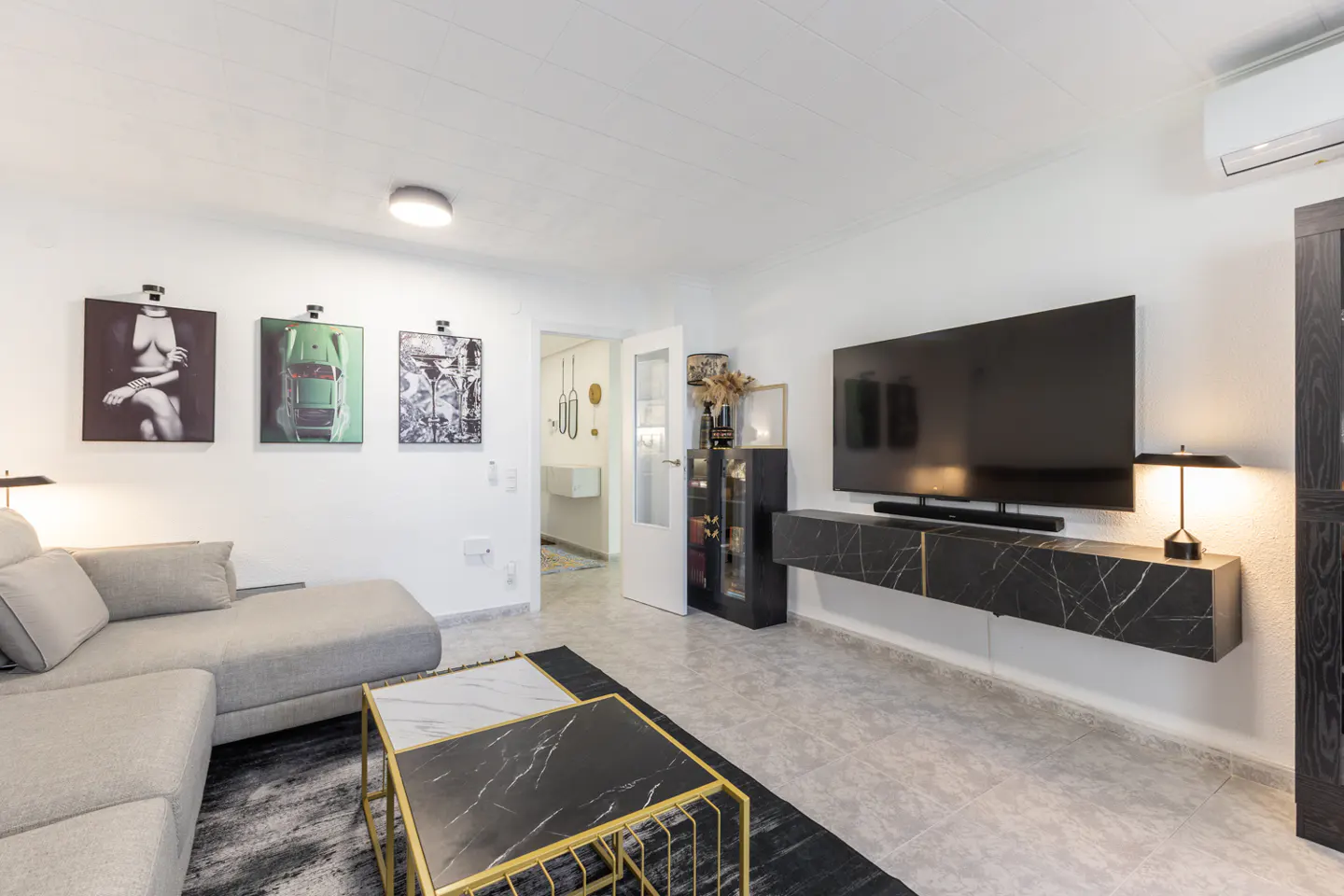 Living room with gray sectional sofa, black and gold coffee tables, and a large TV mounted on a black marble console. Three framed art pieces hang on the wall.