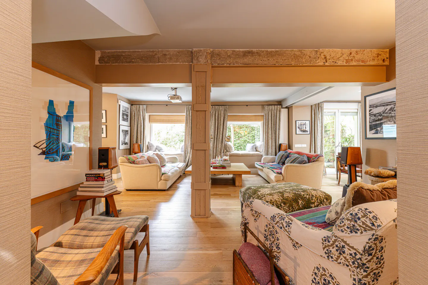 Living room with light wood floors, beige walls, and exposed beams. Two sofas face each other with a wood coffee table between them. Windows with seating areas are behind each sofa.