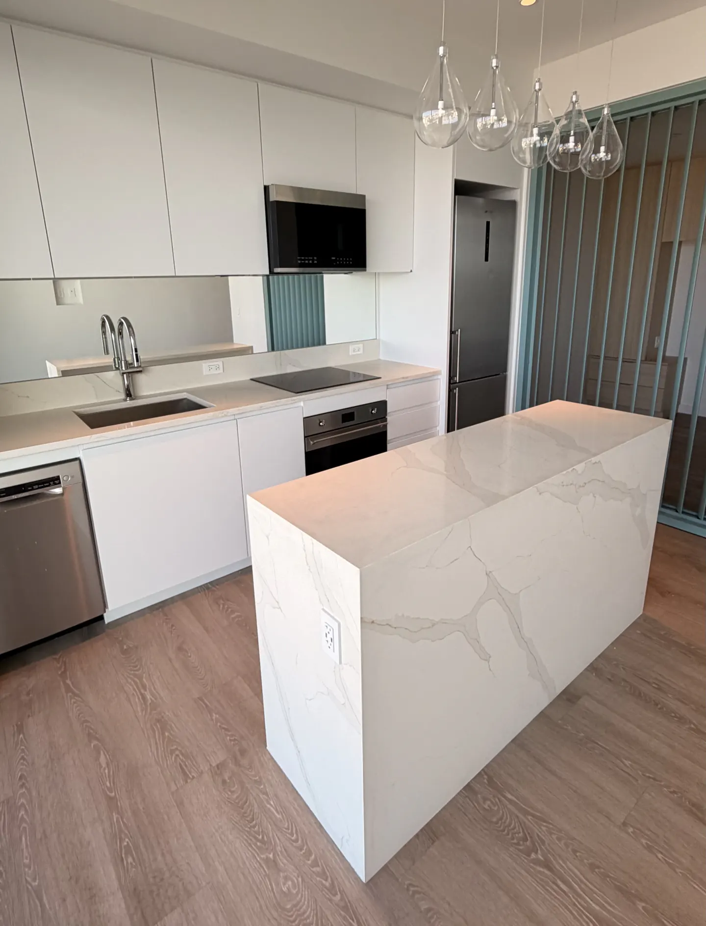 A modern kitchen with white cabinets, stainless steel appliances, and a large marble island. Pendant lights hang above the island.