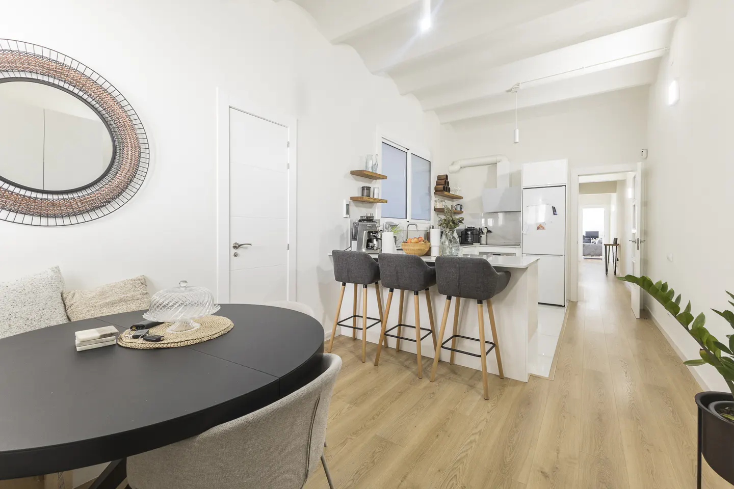 Bright, modern kitchen and dining area with white walls, wood floors, and a black oval table. Three gray bar stools line the kitchen island.