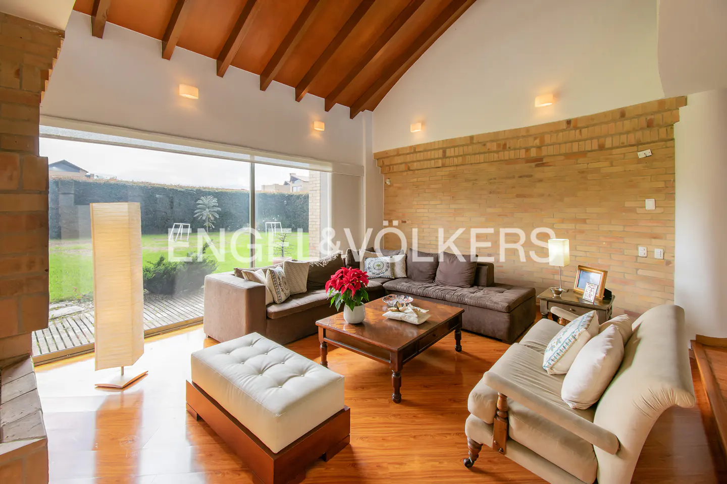 Living room with brown sofas, wood floors, and brick accent wall. Large window overlooks a green lawn. Wooden ceiling beams add warmth.