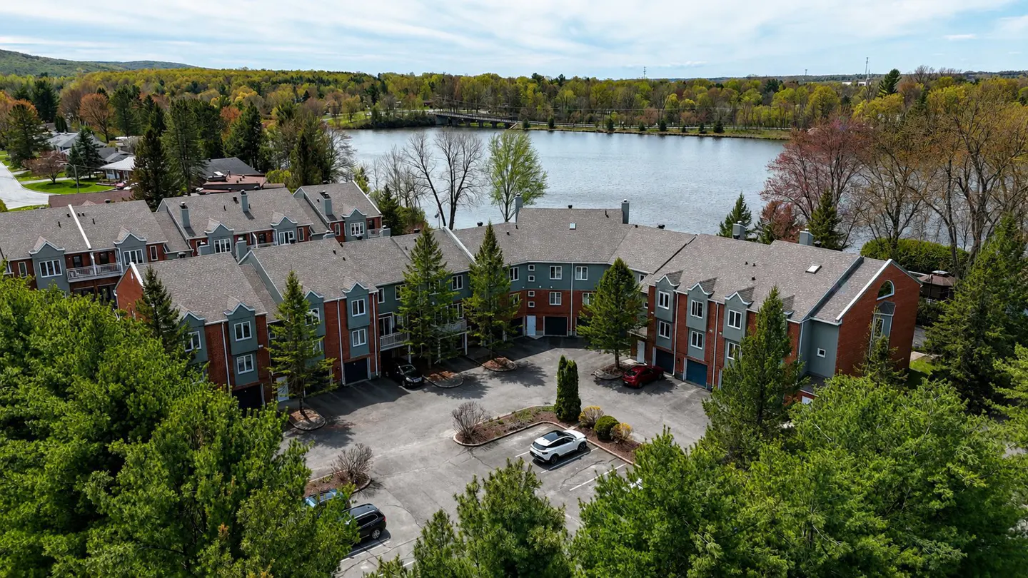 Aerial view of a U-shaped condo complex with a parking lot, surrounded by green trees and a lake in the background.