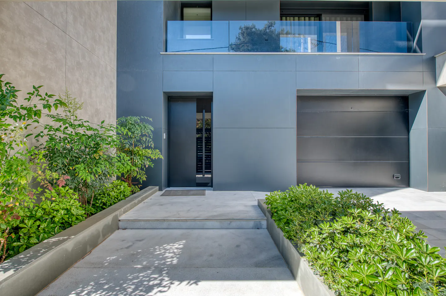 Modern home exterior with gray facade, dark door, and garage. Green plants line the concrete walkway leading to the entrance. Balcony with glass railing above.