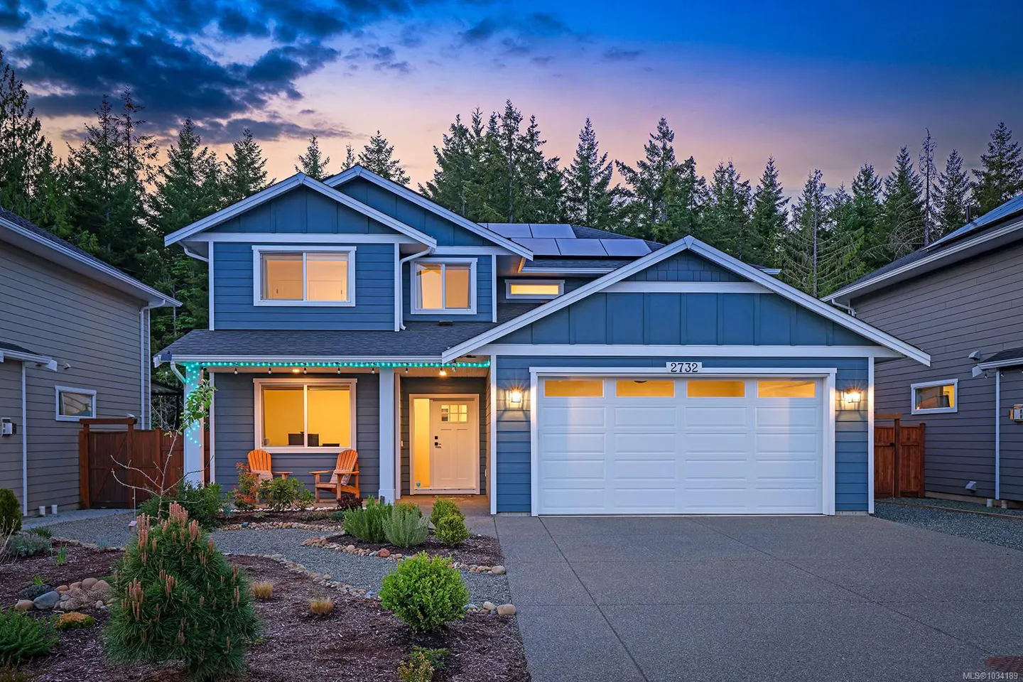 Two-story blue house with white trim, a white garage door, and a white front door. There are solar panels on the roof and a forest in the background.