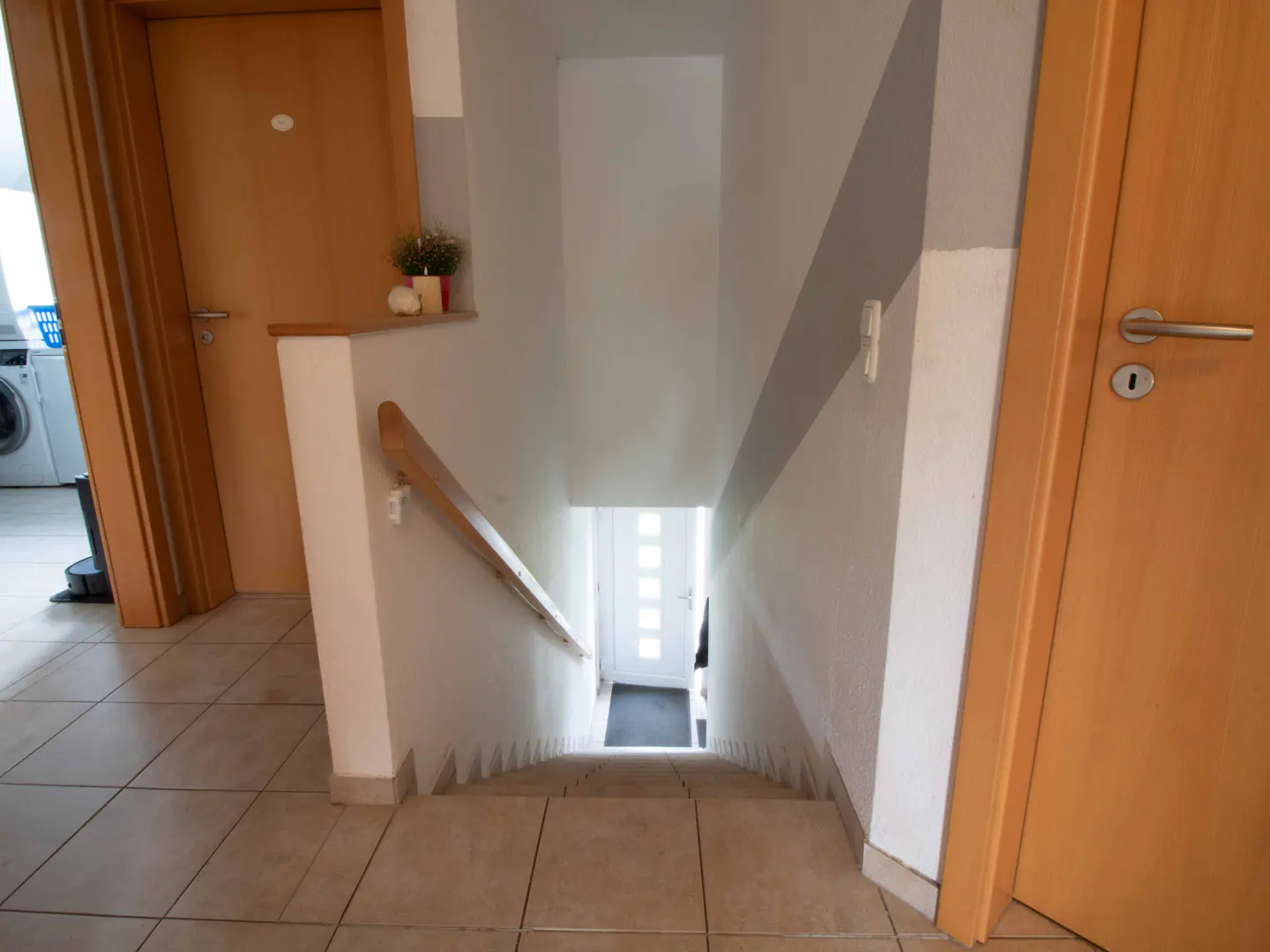 Interior view of a staircase leading down to a white door, with a wooden handrail and beige tile flooring.