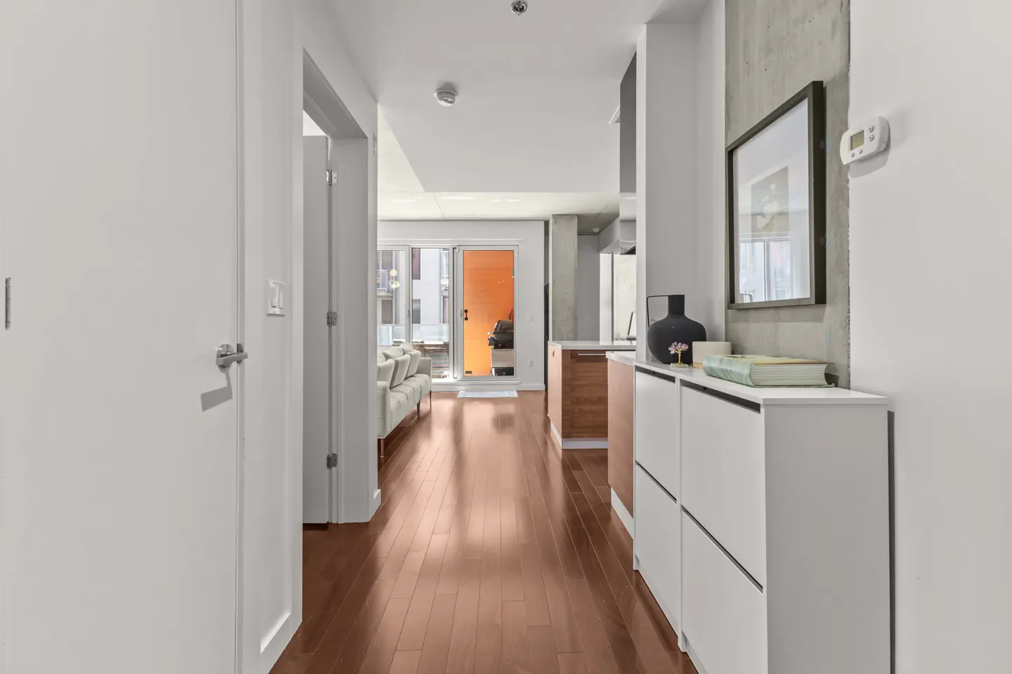 Hallway view with hardwood floors, white walls, and a white cabinet with decor. A framed picture hangs above the cabinet.
