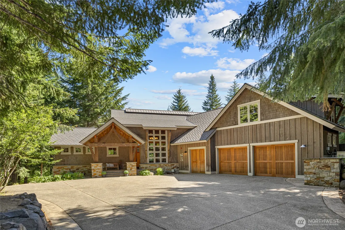 A rustic wood-sided home with a large concrete driveway, two-car garage, and stone accents, surrounded by evergreen trees.