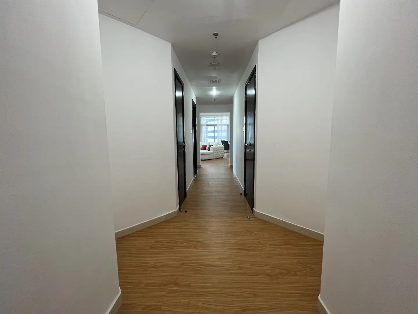 Hallway with light wood floors, white walls, and dark doors. Living room with white sofa visible at the end.