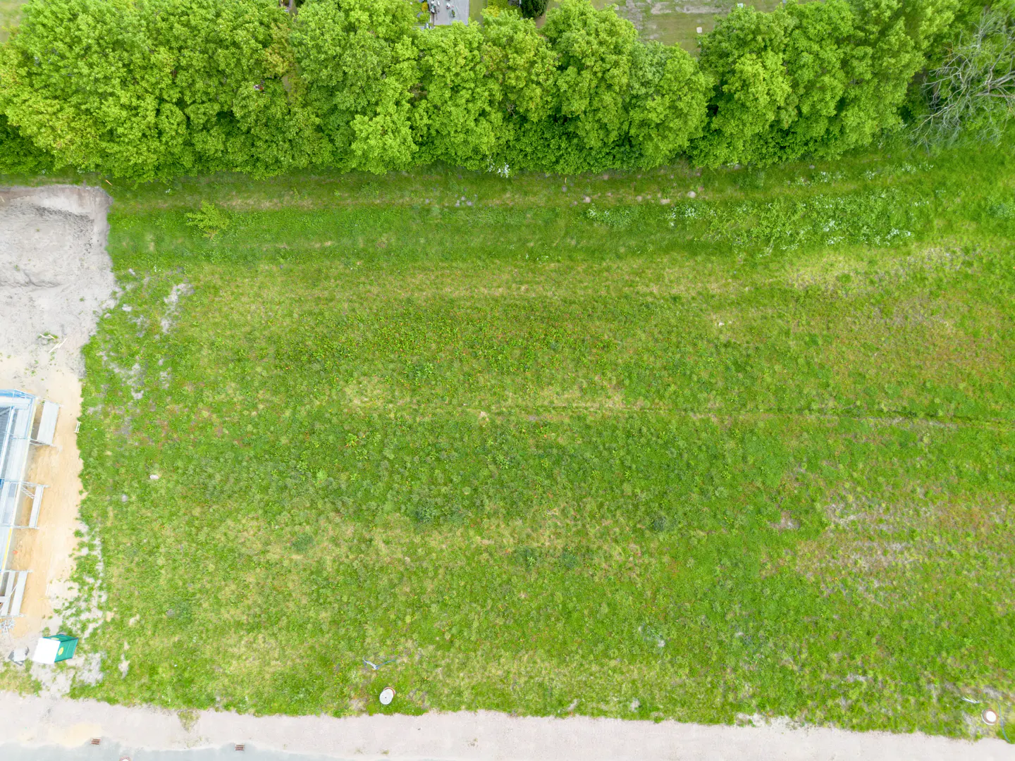 Aerial view of a grassy lot, bordered by green trees at the top and a gray road at the bottom. A building is under construction on the left.