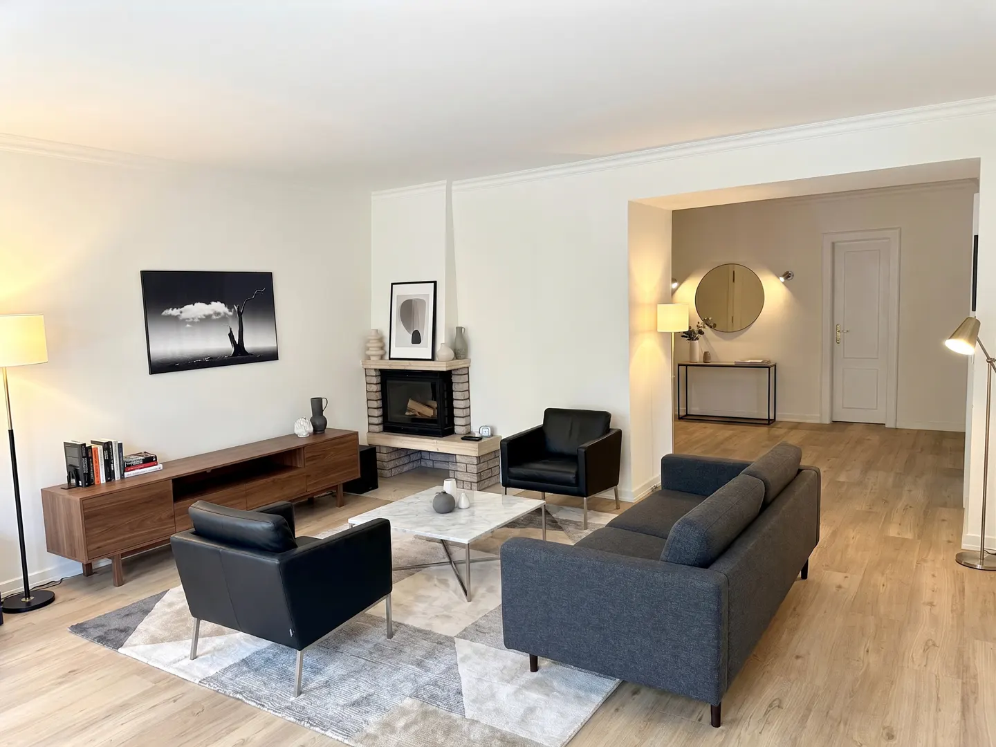 Living room with gray sofa, black chairs, marble table, fireplace, and wood floors. Artwork adorns the walls.