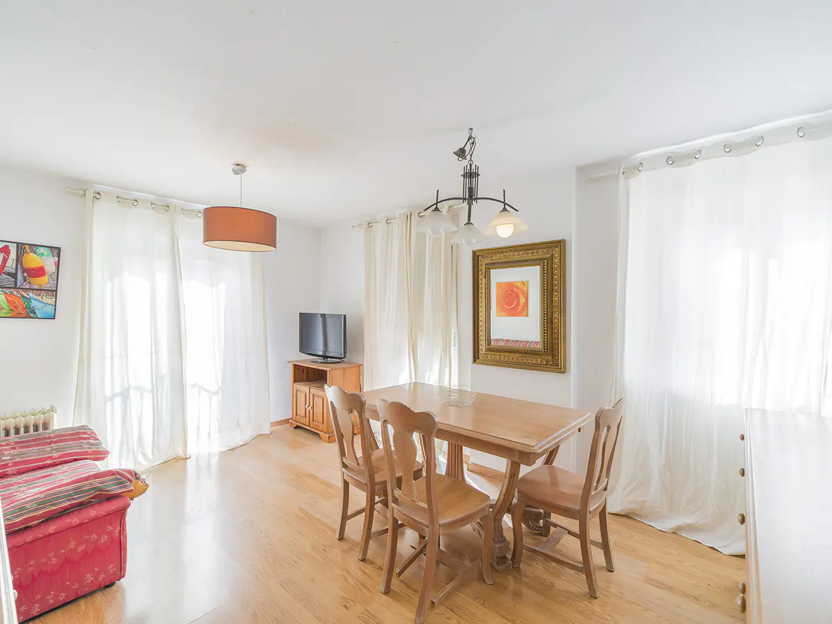 Bright living room with wood floors, a dining table with four chairs, and a red sofa. A TV and framed art add to the homey feel.