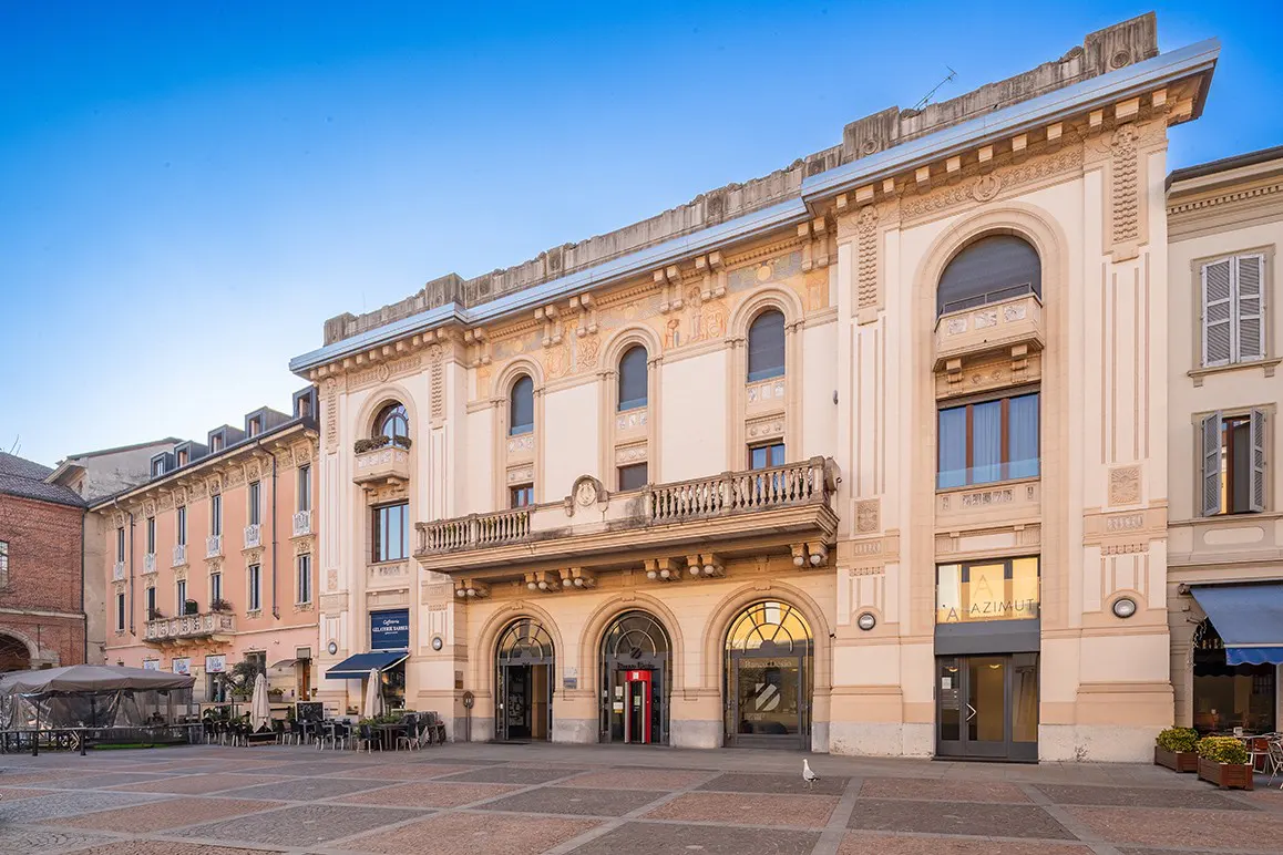 Exterior view of a light beige, ornate building with arched windows and a balcony, on a brick-paved square.