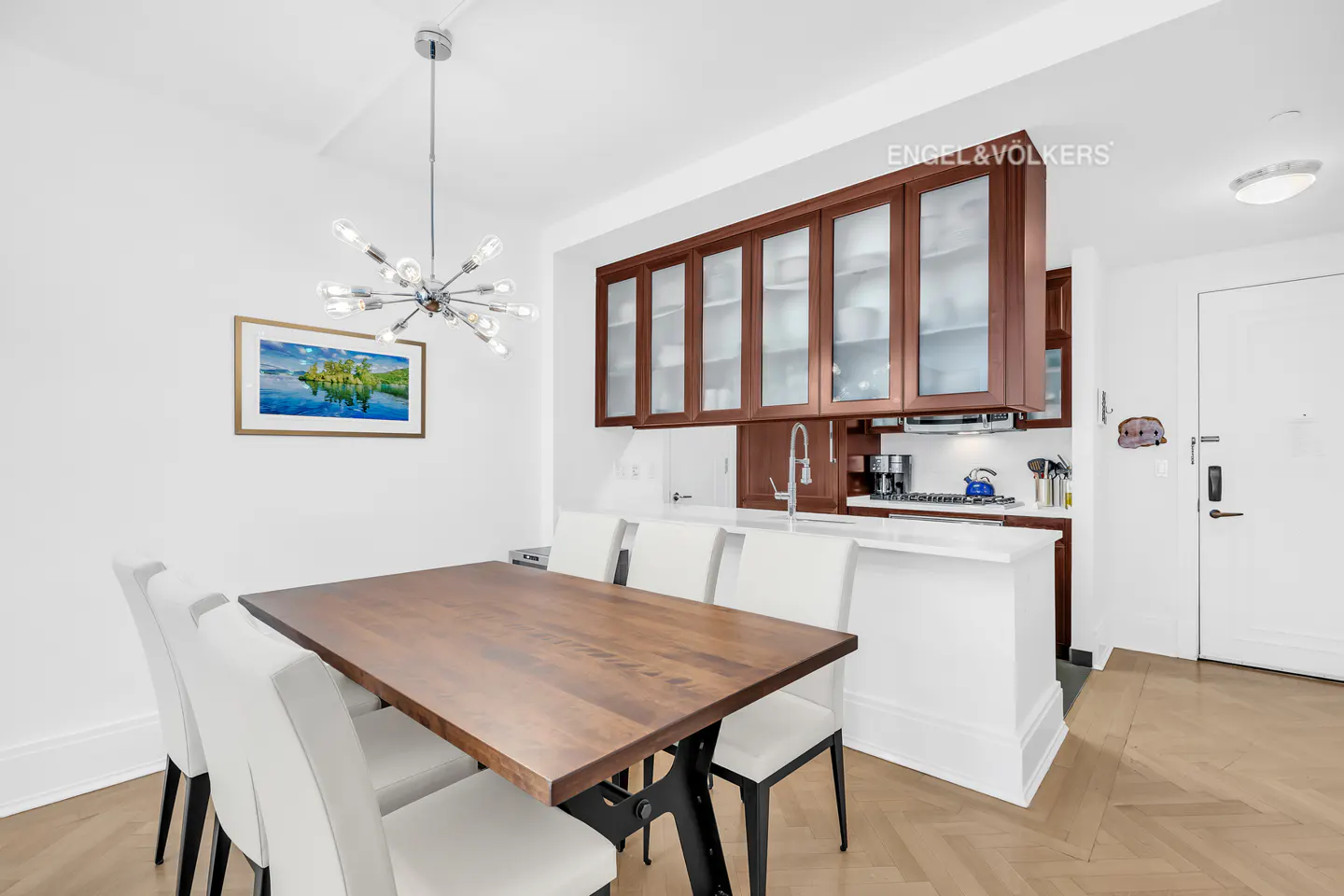 A dining area with a wooden table, white chairs, and a modern chandelier. The kitchen is visible in the background with brown cabinets.