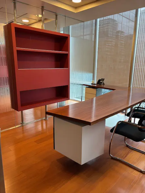 Office interior with a large wooden desk, black chair, and a red bookshelf suspended from the ceiling. The floor is wooden and shiny.
