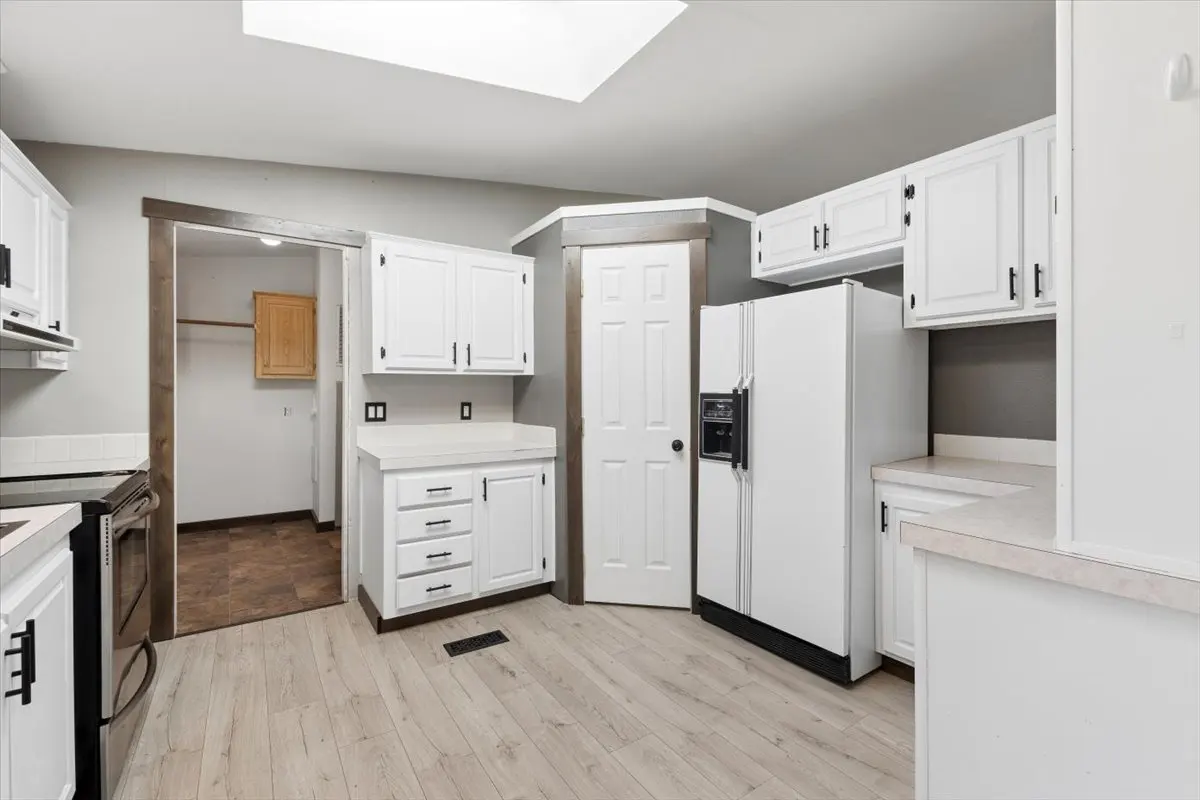 Bright kitchen with white cabinets, appliances, and light wood floors. A doorway leads to another room. Skylight overhead.