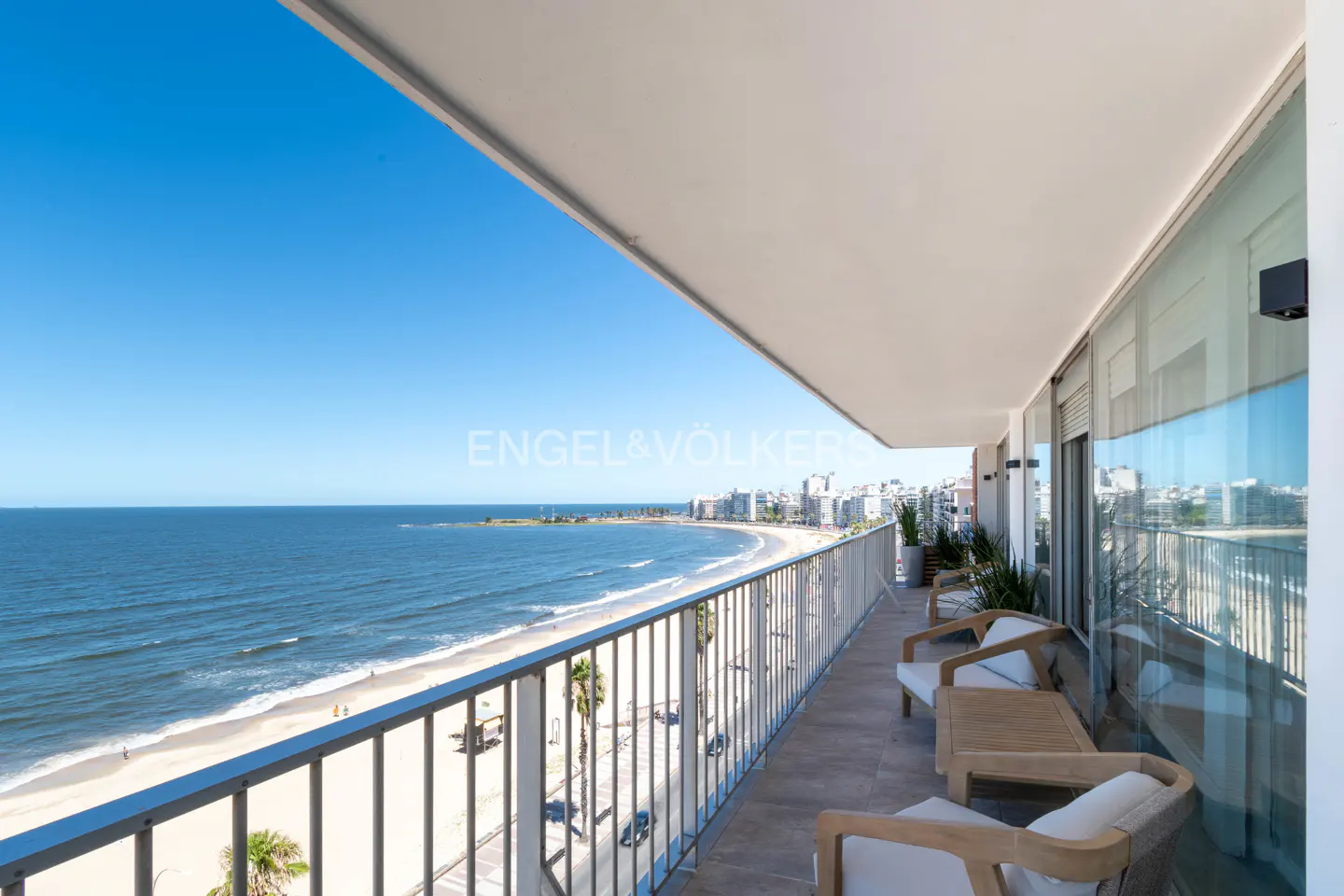 Balcony view of a beach with blue water and sky. Chairs and a table are on the balcony.