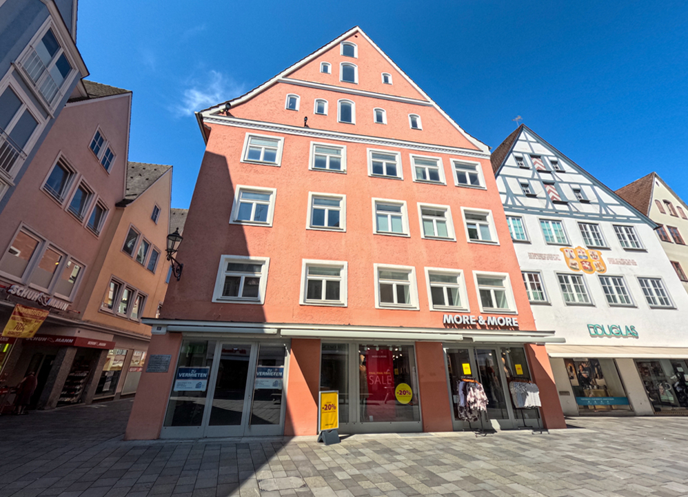 A salmon-colored multi-story commercial building with a peaked roof, featuring a ground-floor storefront on a paved plaza. [AI-generated text]