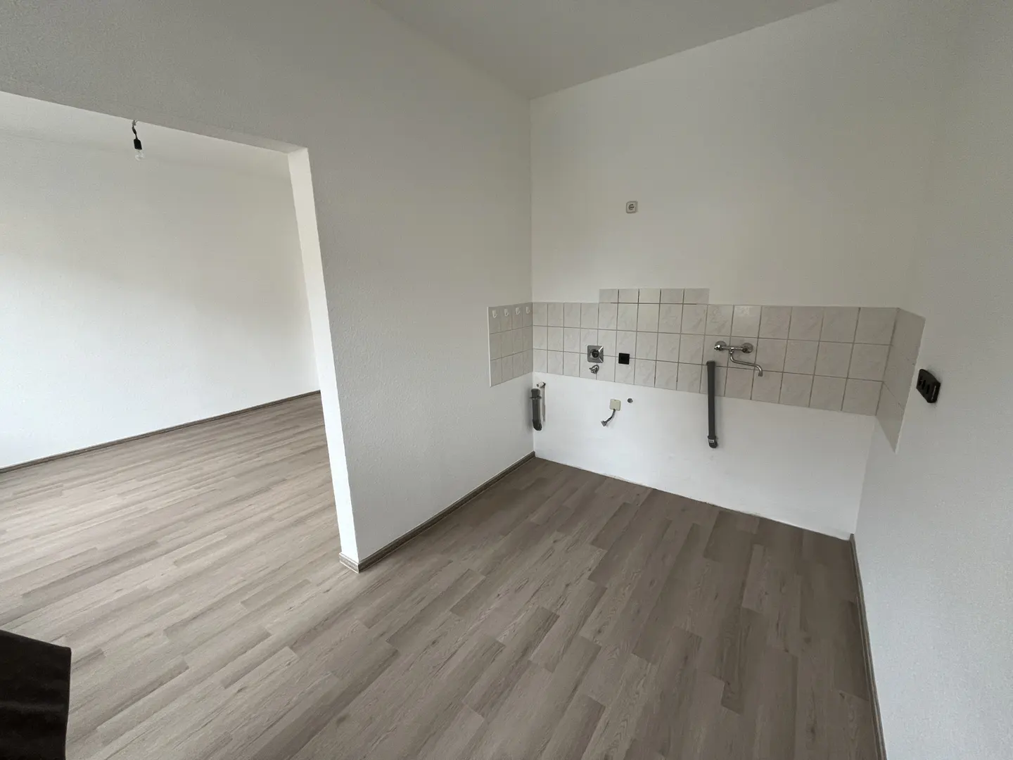 An empty kitchen area with white walls, wood-look flooring, and white tiled backsplash with plumbing fixtures. An open doorway leads to another room.