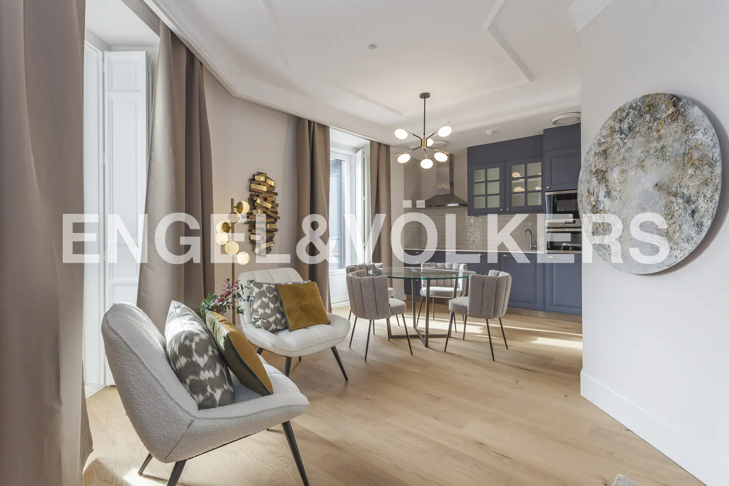 Modern living room with two white armchairs, a glass dining table, and dark blue kitchen cabinets on light wood floors.