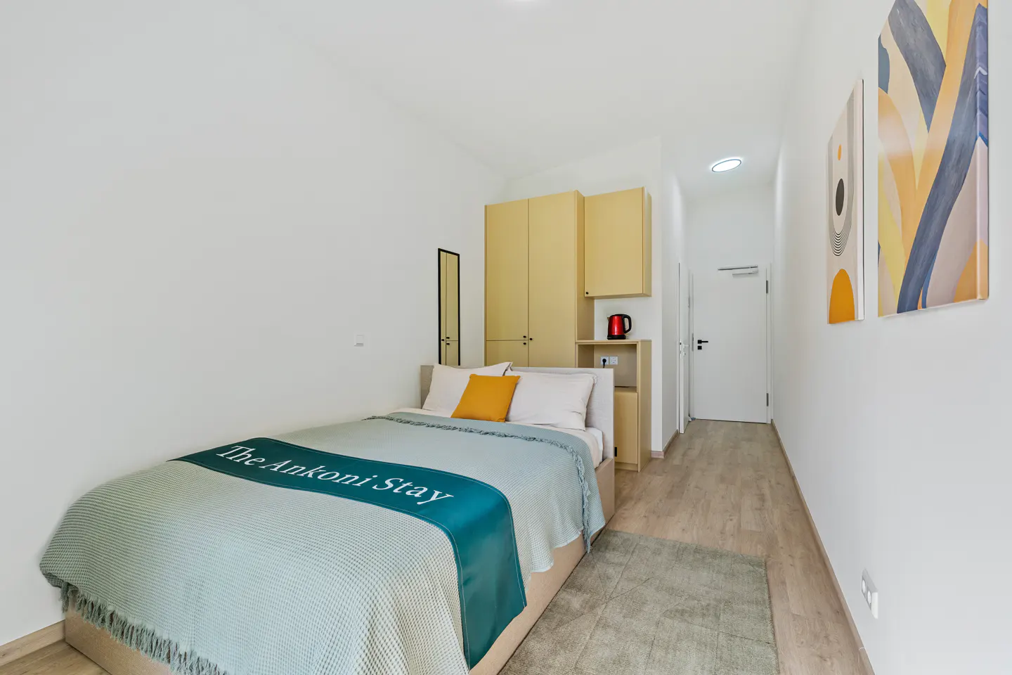 Bright studio apartment with a bed, yellow cabinets, and abstract art on white walls.