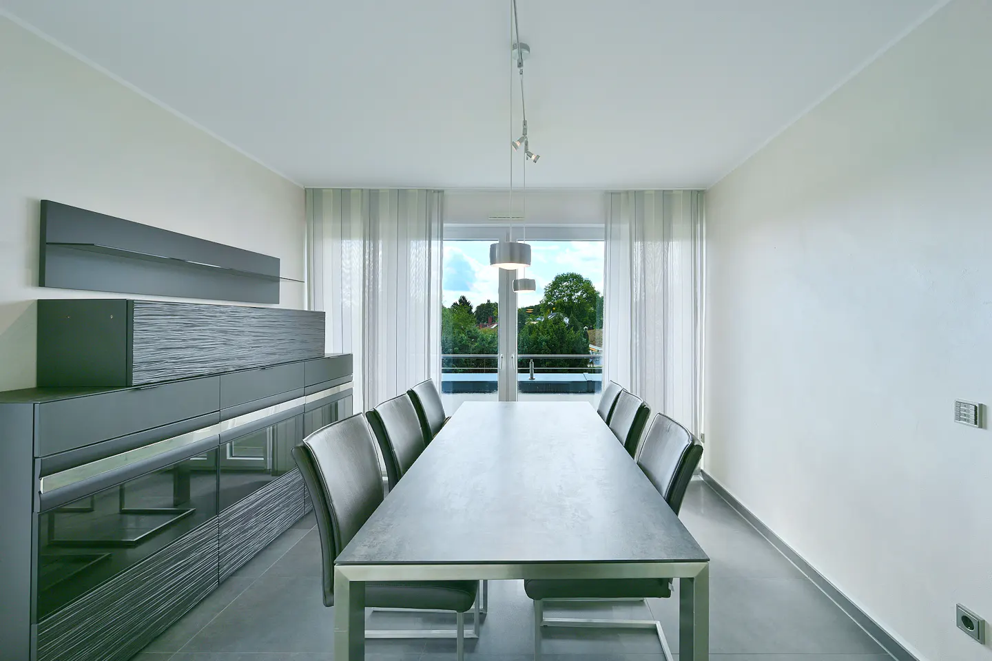 A modern dining room with a long table, gray chairs, and a large window with sheer curtains. A gray cabinet is on the left wall.