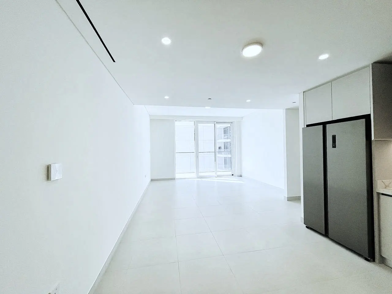 Bright, empty apartment with white walls, tile floor, and sliding glass doors to a balcony. A stainless steel refrigerator is on the right.