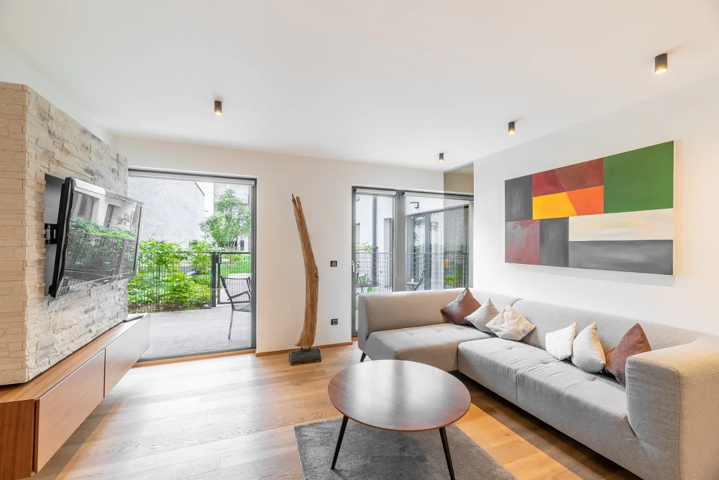 Bright living room with gray sectional sofa, wood floors, and colorful abstract art. Patio visible through open glass doors.