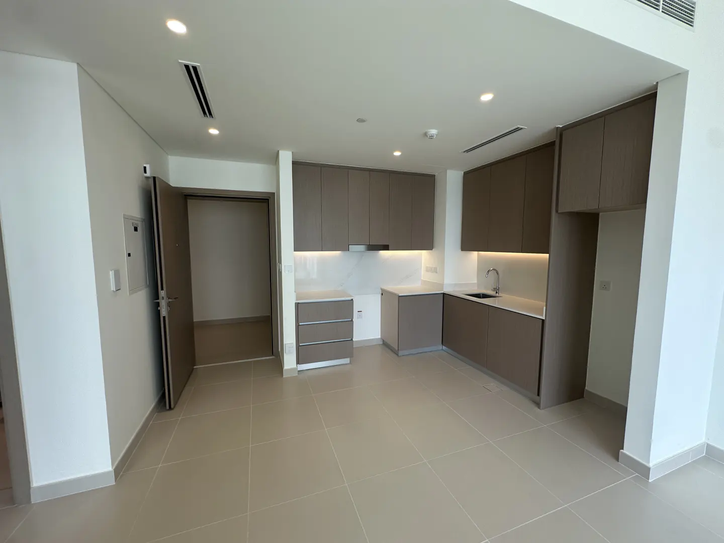 A bright, empty kitchen with beige cabinets, white countertops, and tile flooring. An open doorway is visible on the left.