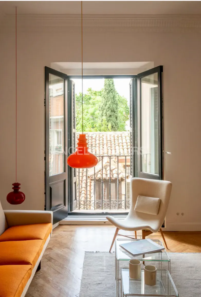 Bright living room with orange sofa, beige chair, and glass tables. Open doors reveal a balcony with a view of trees and rooftops.