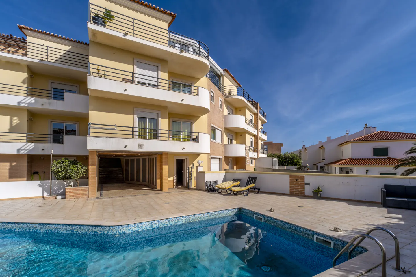 Exterior view of a yellow apartment building with balconies, a pool, and lounge chairs.