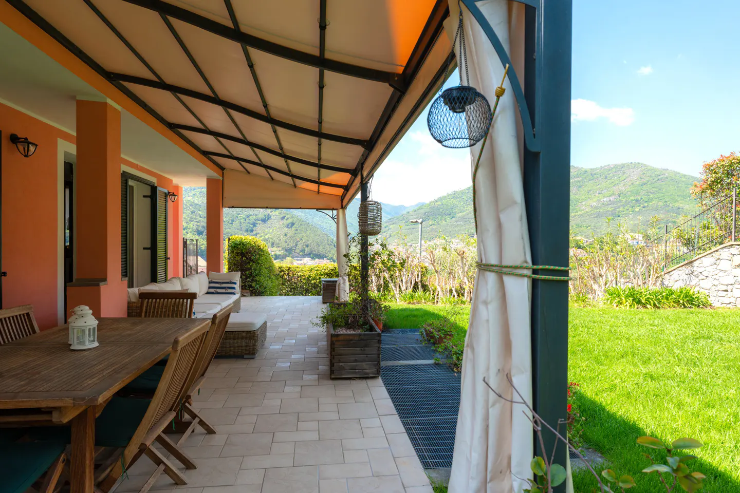 Covered patio with wood table and chairs, white sofa, and mountain view. Peach-colored house with black shutters. Green lawn.