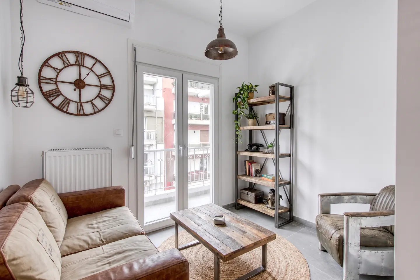 Living room with brown leather sofa, wood coffee table on jute rug, metal bookshelf, and large clock on white wall.
