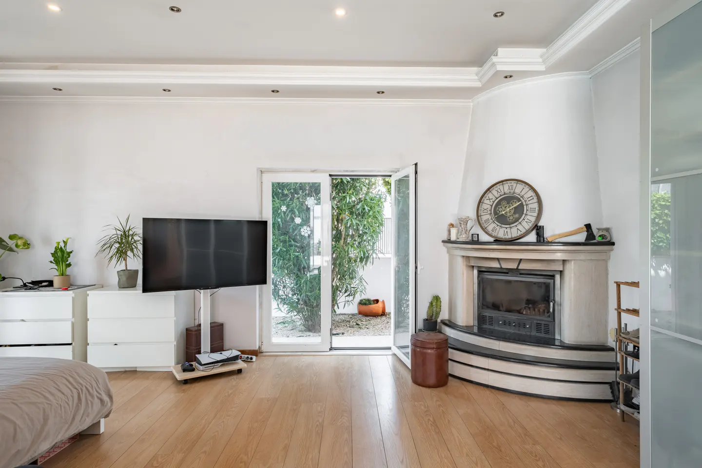 Bright living room with wood floors, white walls, and a fireplace. A TV stands near open doors to a green outdoor space.