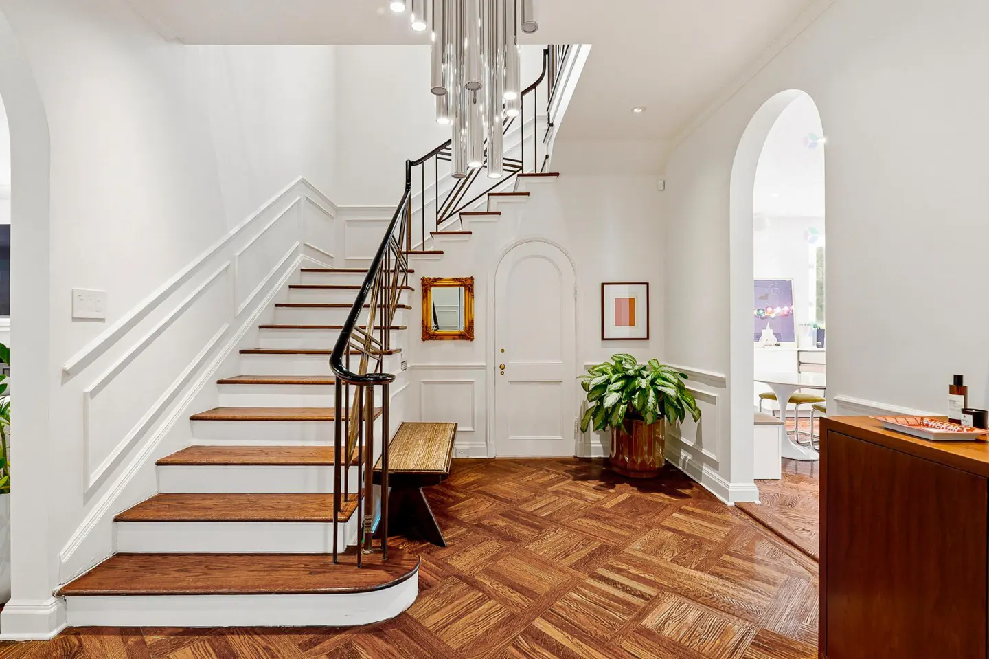 Bright foyer with wood floors, white walls, and a staircase with black railing. A bench sits under a gold-framed mirror. A plant adds a touch of green.
