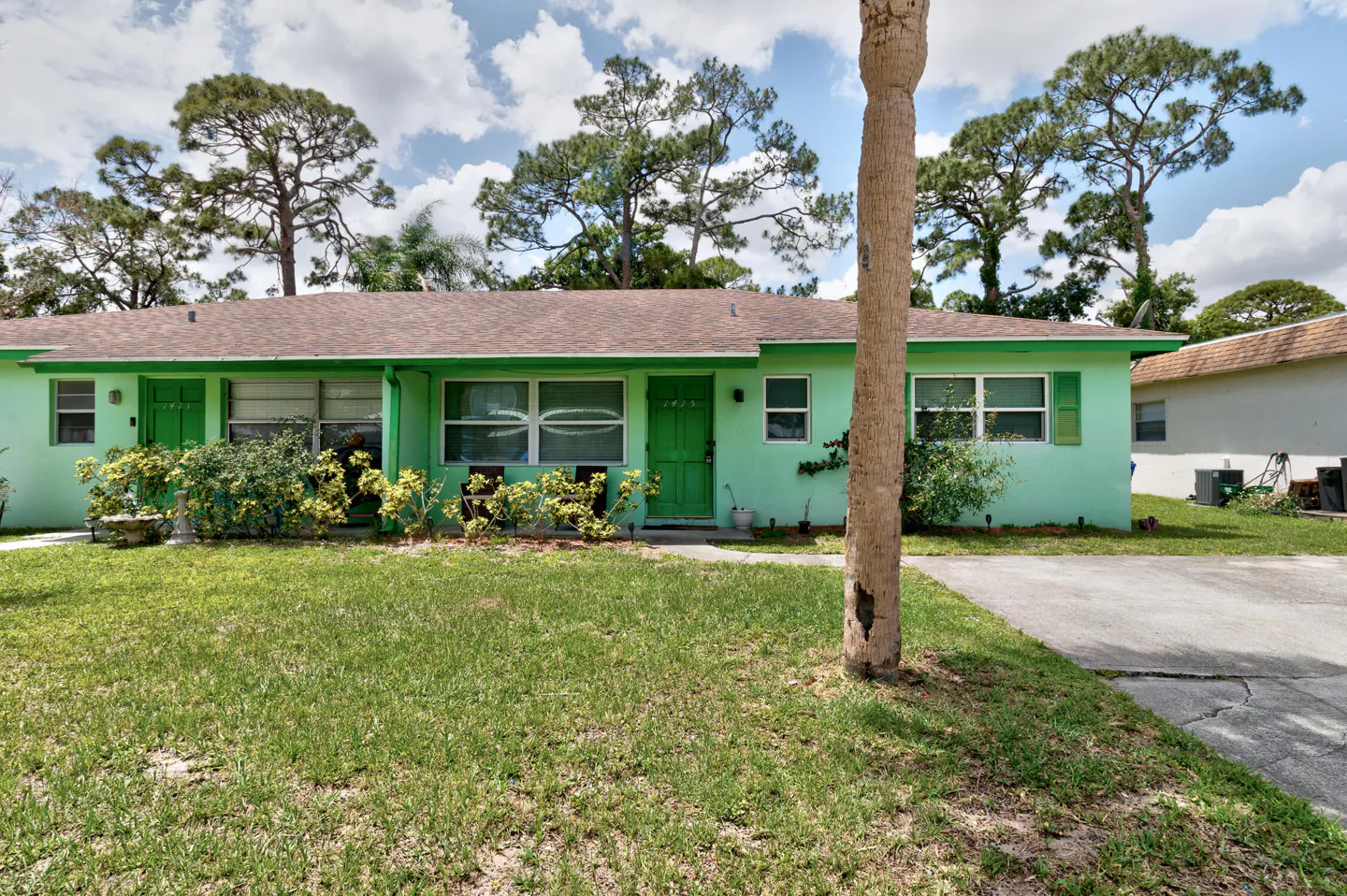 Exterior of a light green single-story house with a brown roof and green door, surrounded by a green lawn and trees.