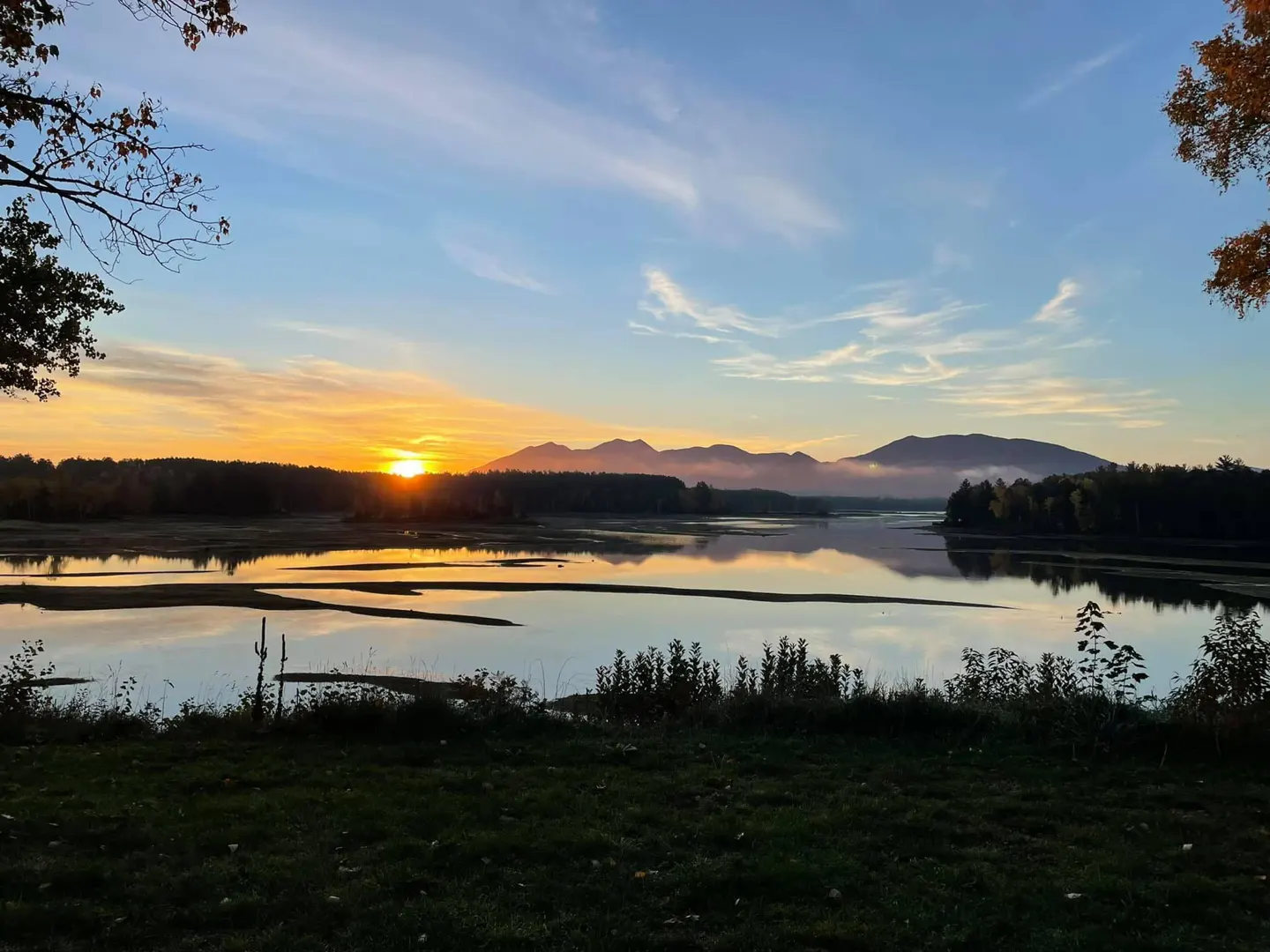 Sunrise over a calm river, reflecting orange light. Mountains are visible in the distance, with trees lining the riverbanks.