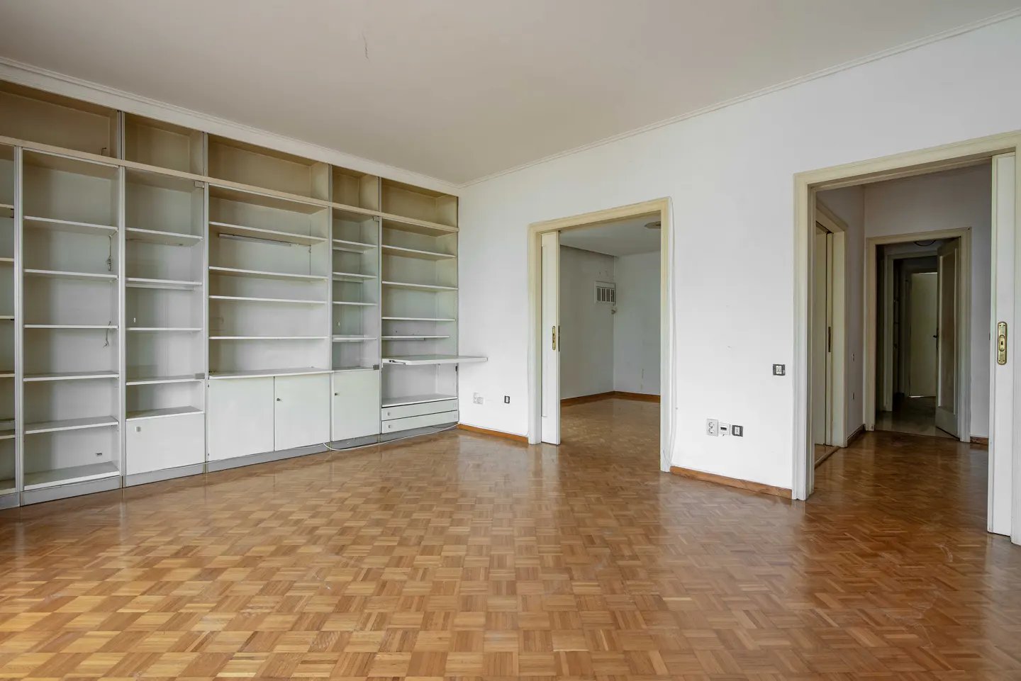 Empty room with parquet floor, white walls, and a large built-in white bookcase. Two doorways lead to other rooms.