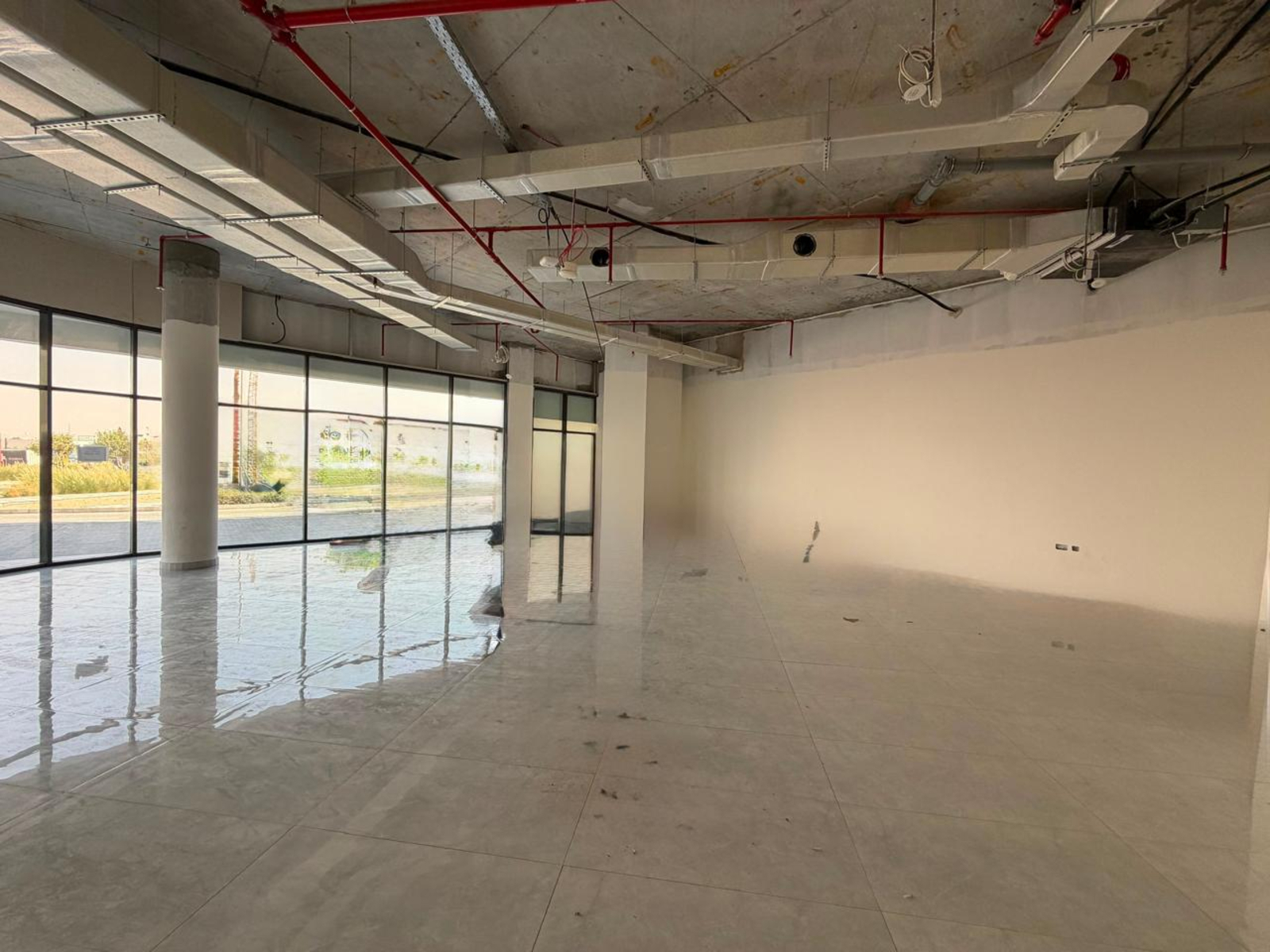 An unfinished commercial space with polished tile floors, floor-to-ceiling windows, and exposed industrial ceiling pipes.