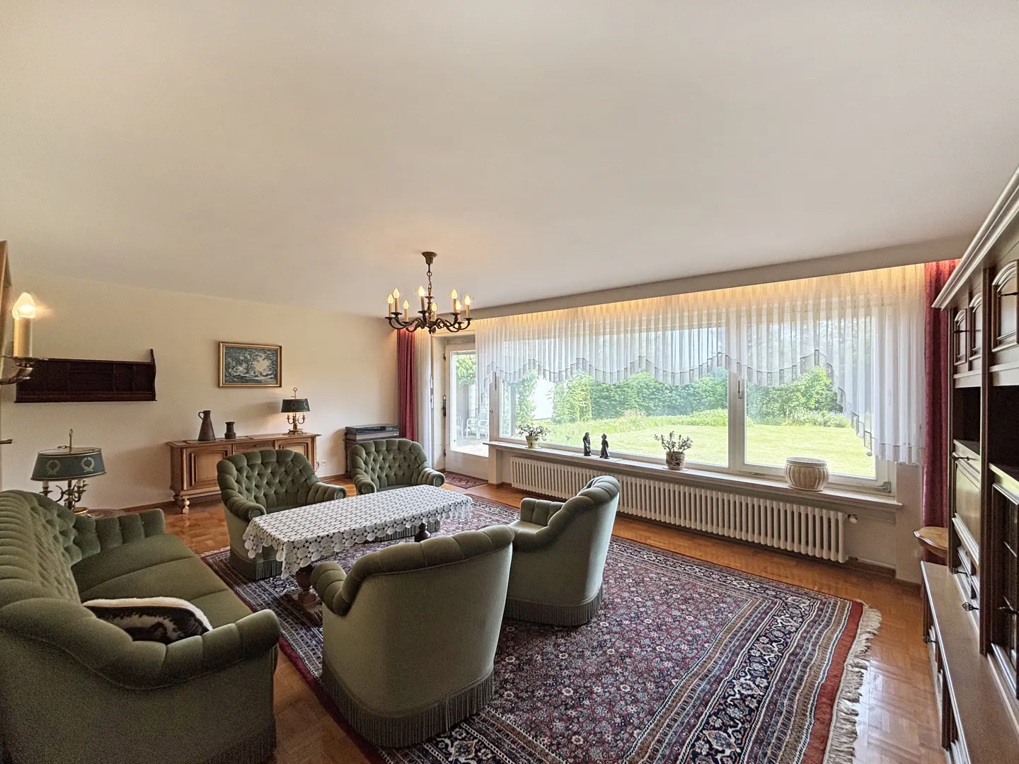 A living room with green furniture, a patterned rug, and a large window with white curtains.