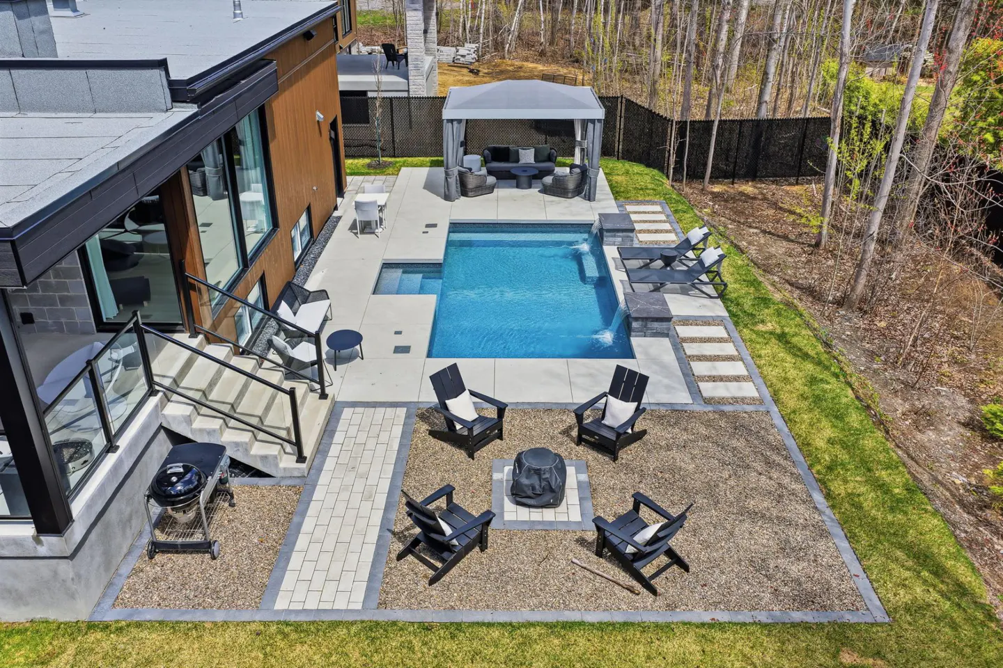 Aerial view of a modern backyard with a blue pool, gray gazebo, fire pit with black chairs, and a grill near the house.