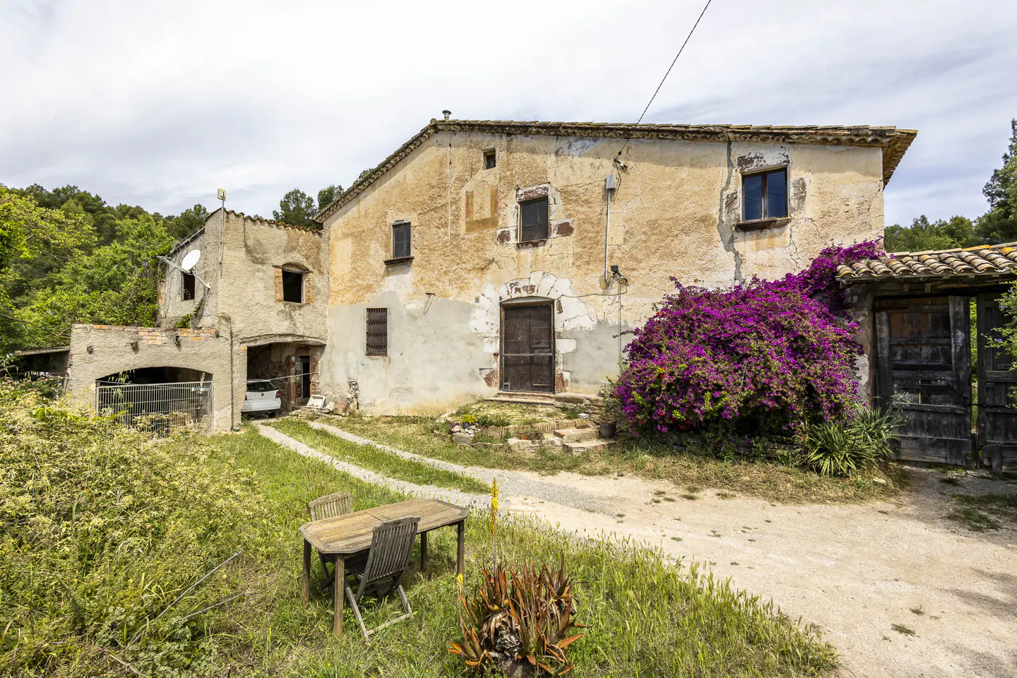 Exterior of a two-story, tan stucco farmhouse with a dark wood door and purple bougainvillea. A wooden table and chairs sit on the lawn.