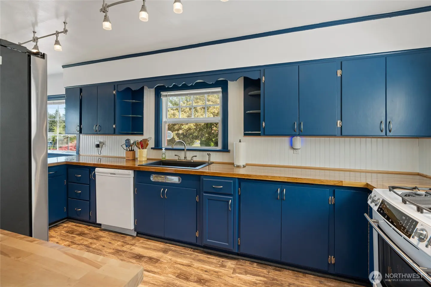 A kitchen with blue cabinets, wood countertops, and wood-look flooring. A window is above the sink. Track lighting is on the ceiling.