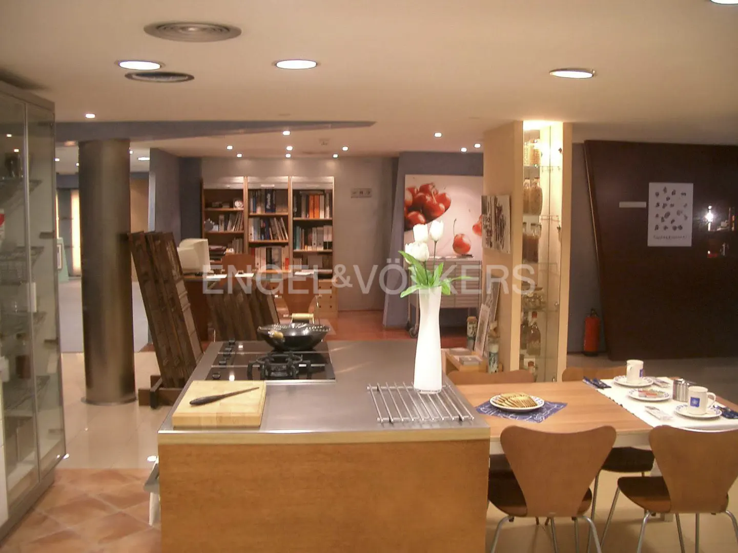 A modern kitchen with a stainless steel island, wooden cabinets, and a dining table set for a meal. A bookshelf and cherry art are in the background.