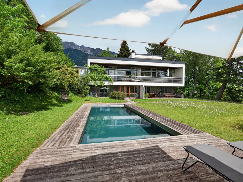 Modern two-story house with a pool and wooden deck. Green lawn, trees, and mountains in the background. Blue sky with white clouds.