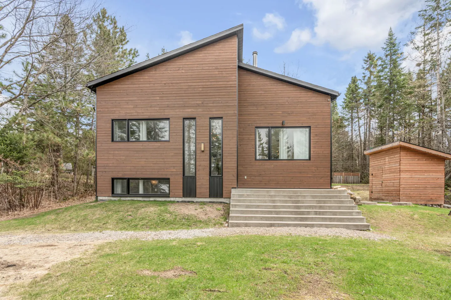 Modern brown wood home with black trim, concrete steps, and a small shed in a grassy yard with trees.
