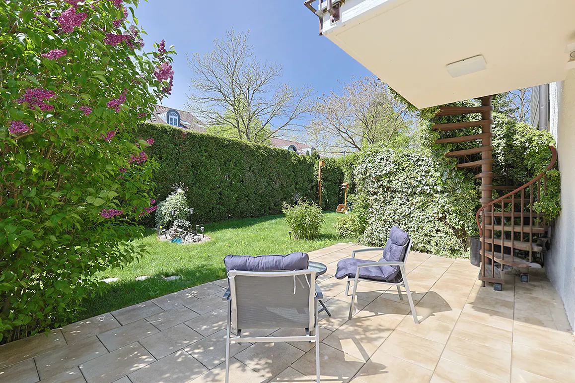 Patio with two chairs, a small table, and a spiral staircase. Green lawn and bushes in the background.